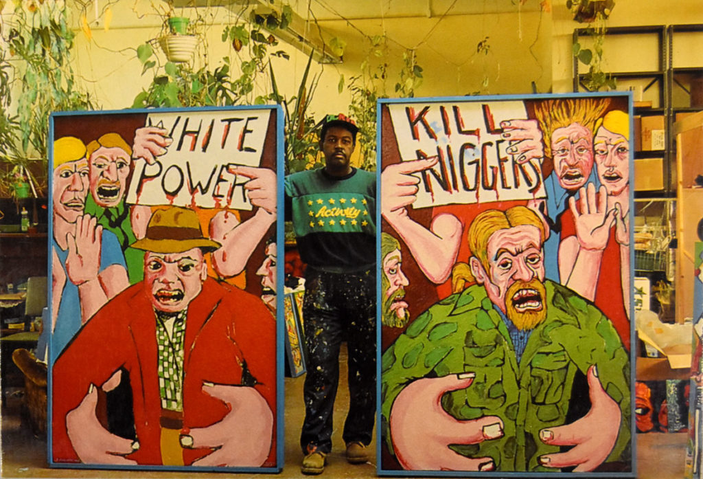 Dana Chandler photographed in his studio for the postcard for his "The more things change, the more things remain the same, Let My People Go!" retrospective at Massachusetts College of Art, Feb. 23 to March 20, 1987. On the left is his painting "The Beast," 1967-68, and on the right is "The Beast Revisited-Forsyth County, Georgia," 1987.
