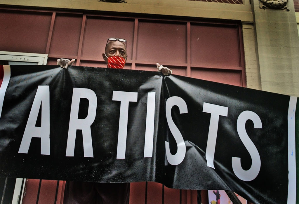 Jeff Chandler--brother of artist, teacher and activist Dana Chandler who founded the African American Master Artists In Residence Program in 1978--holds a banner as artists and supporters of the program protest outside the studios building at 76 Atherton St. in Boston's Jamaica Plain neighborhood, June 27, 2020. (Photo © copyright Don West / fOTOGRAfIKS)