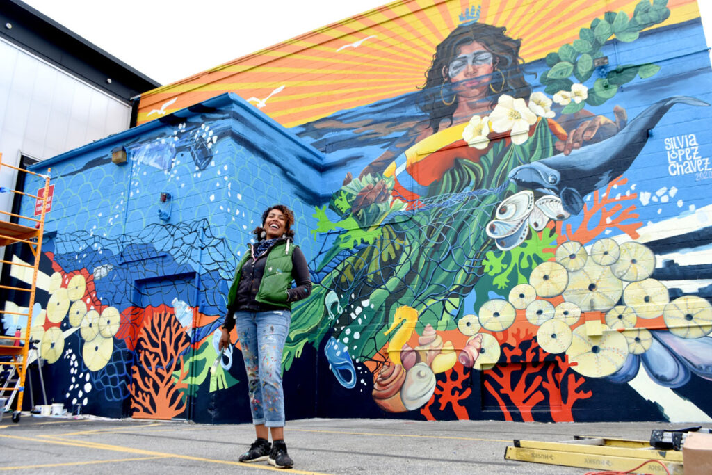 Silvia Lopez Chavez with her mural for "Sea Walls: Artists for Oceans, Boston 2020," from PangeaSeed Foundation in collaboration with HarborArts at the Boston Harbor Shipyard in East Boston, Sept. 18, 2020. (© Greg Cook photo)