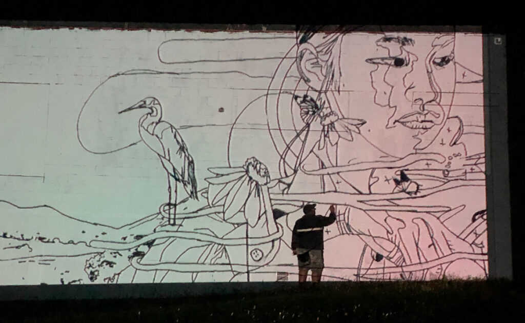 Cedric "Vise1" Douglas using a projection to trace the sketch onto the wall for his mural at the former Medfield State Hospital, May 2021. (Courtesy Cultural Alliance of Medfield)