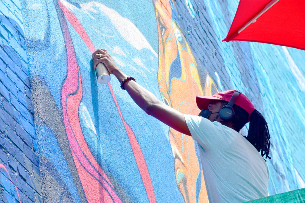 Taj Francis of Jamaica paints at 150 Liverpool St., East Boston, for the "Sea Walls" mural project, July 23, 2021. (©Greg Cook photo)
