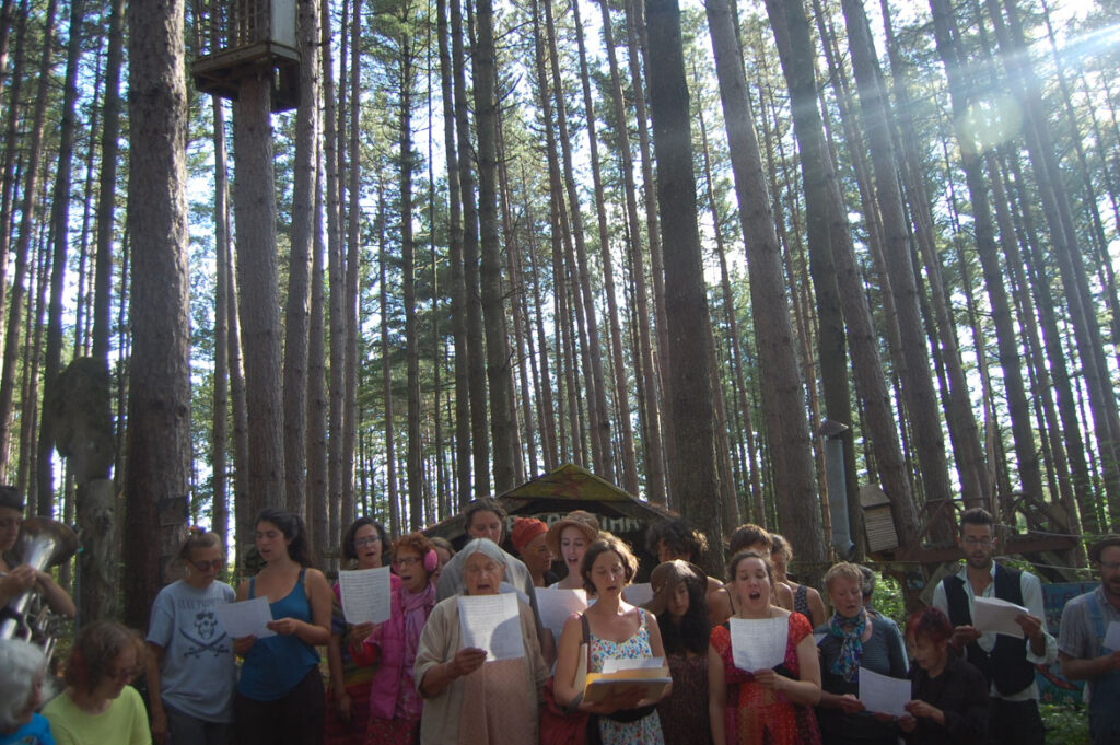 Elka Schumann (center) sings during a memorial in the pine forest at Bread and Puppet, Glover, Vermont, Aug. 21 2015. (©Greg Cook photo)