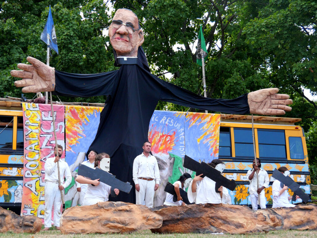 Bread and Puppet Theater performs an act honoring Archbishop Oscar Romero who was murderd in 1980 in El Salvador. From its “Apocalypse Defiance Circus” on Cambridge Common, Sept. 4, 2022. (© Greg Cook photo)
