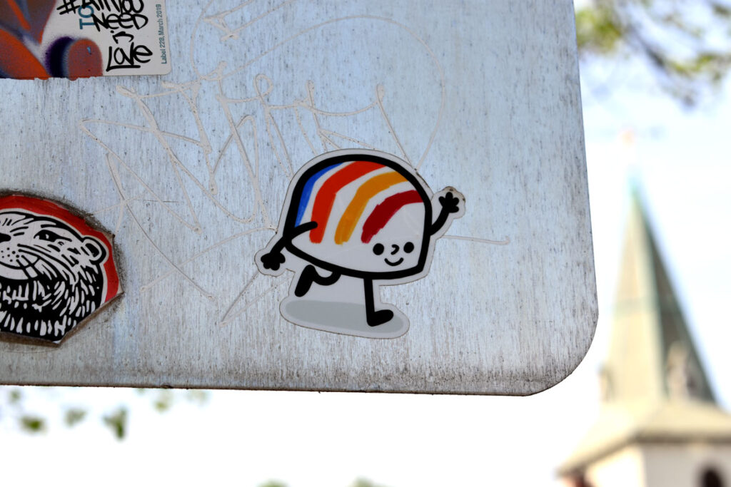 Street art sticker spotted on the back of a sign on Broadway by Cambridge's Sennot Park, riffing on Corita Kent's iconic 1971 rainbow gas tank design in Boston's Dorchester neighborhood. May 3, 2021. (©Greg Cook photo)