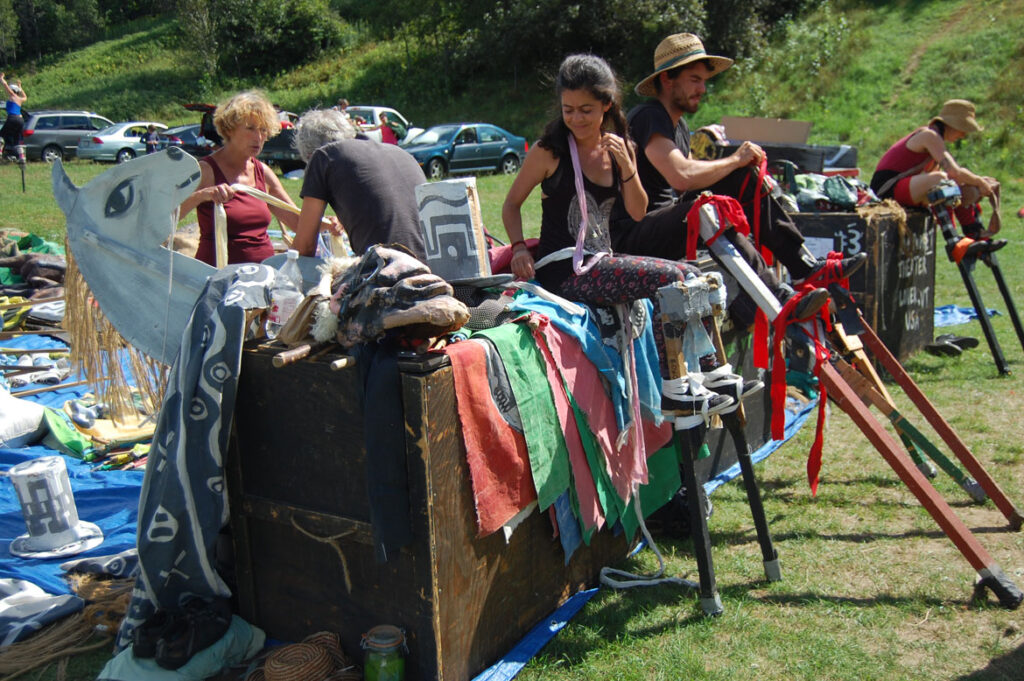 Genevieve Yeuillaz (background, left) helps her husband Remy Paillard strap into stilts for a Bread and Puppet circus rehearsal, Glover, Vermont, Aug. 22, 2015. (©Greg Cook photo)