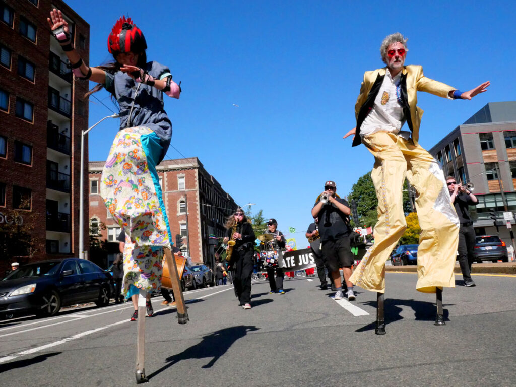 Stilters dance in front of the Dirty Water Brass Band in the Honk parade from Somerville's Davis Square to Cambridge's Harvard Square, Sunday, Oct 9, 2022 (©Greg Cook photo)