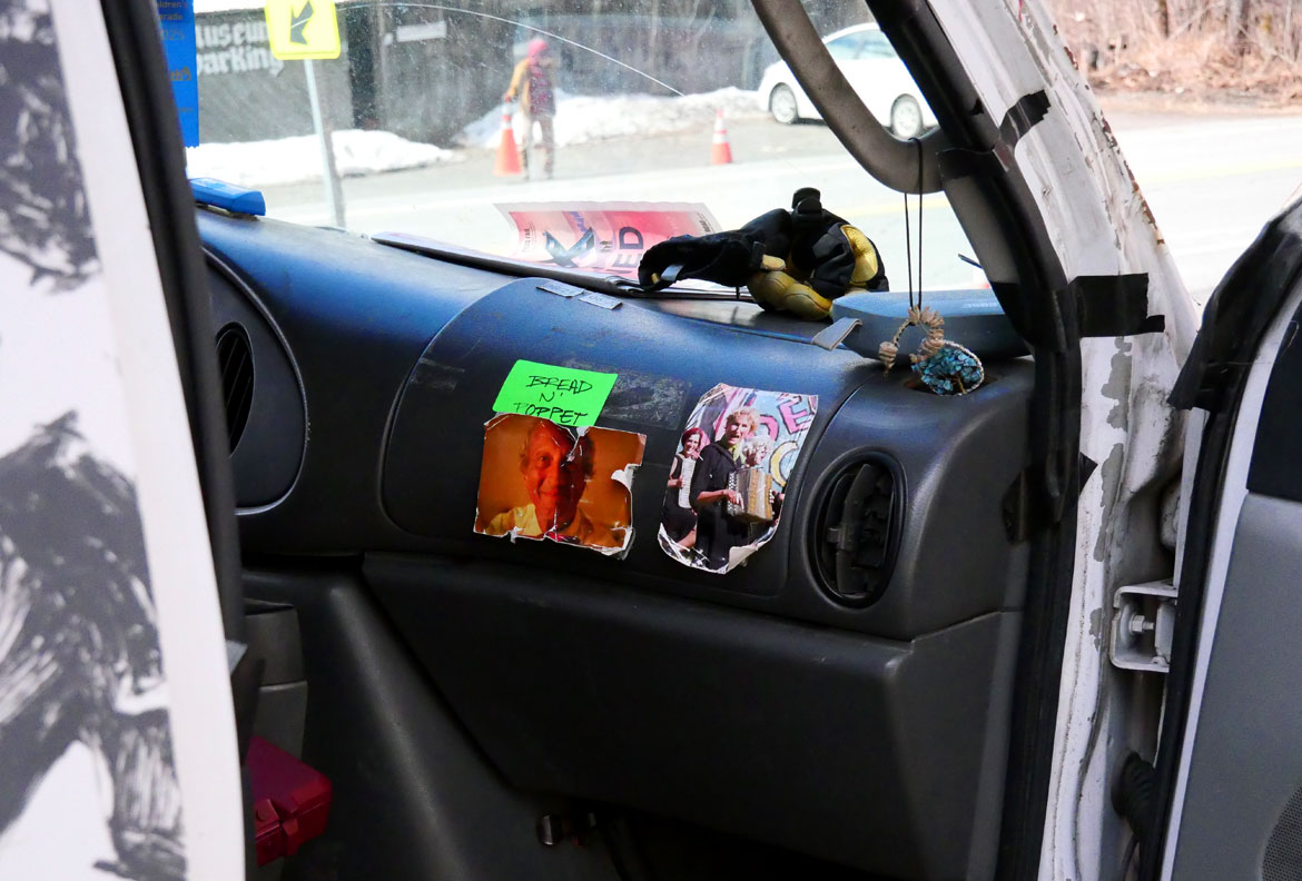Photos of the late Remi Paillard and Genevieve Yeuillaz, a couple who long worked with Bread and Puppet Theater, taped to the dashboard of the company's black and white van, Glover, Vermont, April 11, 2026. (©Greg Cook photo)