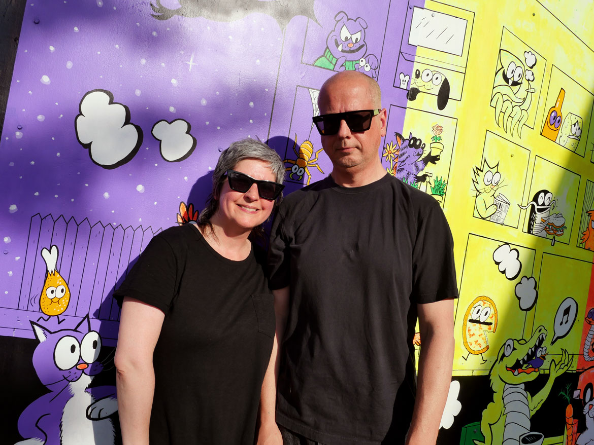 Blipblipbeep (Jennifer Puriņš, left) and “Ansis Puriņš in front of their mural "All This, All at Once" outside the Harvard Ed Portal in Allston, June 2026. (Greg Cook photo)