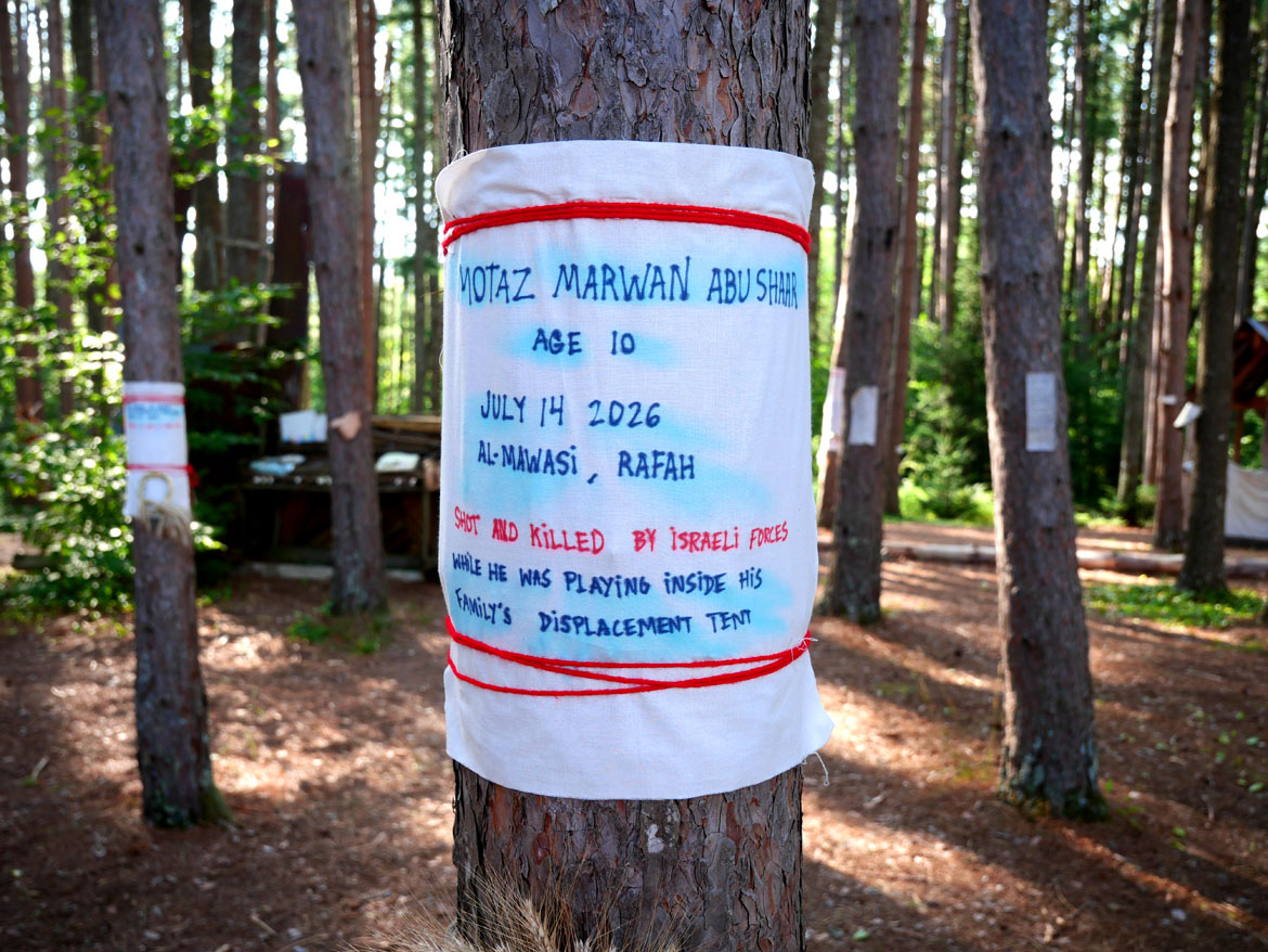 Memorials of killed Palestinians and Iranians tied to trees in the Pine Forest for Bread and Puppet Theater's "Upside Down World Resurrection Pageant," performed on their Glover, Vermont, farm, July 26, 2026. (©Greg Cook photo)